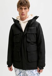 Sort vandtæt jakke med fire store frontlommer, hætte og justerbare ærmer. Har et logo på brystet og en glat tekstur.