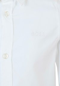 Camicetta bianca con bottoni, realizzata in tessuto morbido, caratterizzata da colletto classico, bottoni trasparenti e logo impresso sul petto.