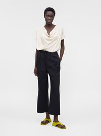 Femme portant un haut drapé crème à manches courtes, un pantalon noir taille haute à jambes larges et des sandales ouvertes jaunes vives, debout avec une main dans la poche.