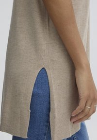 Beżowy sweter dzianinowy z bocznymi rozcięciami i ściągaczem. Stylizowany z niebieskim dżinsami, podkreślający gładką fakturę i swobodny fason.
