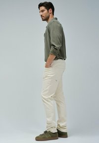 Hombre con camisa de botones verde oliva, pantalones color crema y zapatillas verde oliva, de pie de perfil con las manos en los bolsillos.