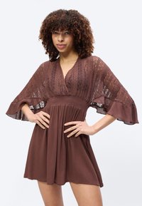 Γυναίκα με σγουρά μαλλιά που φοράει καφέ δαντελένιο και υφασμάτινο φόρεμα με φαρδιά μανίκια, στέκεται με τα χέρια στη μέση μπροστά σε απλό φόντο.