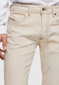 Jeans de denim color beige con salpicaduras en azul claro, diseño de cinco bolsillos, cierre de botón metálico y detalles de costura en contraste.