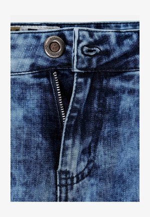 Jean en denim dans un délavage bleu moyen, avec une fermeture éclair, un bouton en cuivre et des détails cousus autour des poches et de la taille.
