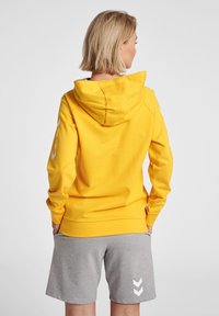 Gul hoodie med en framficka och huva, tillverkad av mjukt tyg; parat med grå shorts som har ett vitt chevronmönster på sidan.