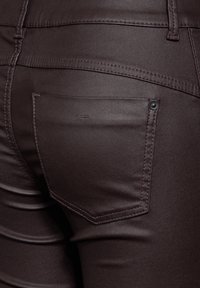 Bruna denimbyxor med en bakfick med en svart knapp, subtil sömnad och en slät, något blank yta.