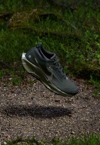 Tumši zaļš sporta apavs ar redzamu Nike logo, karājas virs grants ceļa ar zaļu lapu fonā.