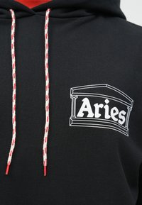 Mustat hupparit, joissa on valkoiset ja punaiset narut, sekä valkoinen graafinen kuva klassisesta rakennuksesta ja "Aries"-teksti rinnassa.