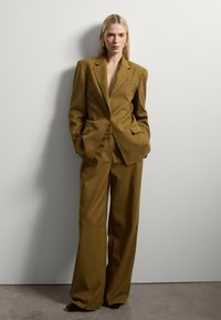 Traje marrón a medida con un blazer de escote en V profundo y pantalones de pierna ancha. Presenta hombros estructurados y bolsillos frontales. Textura suave.