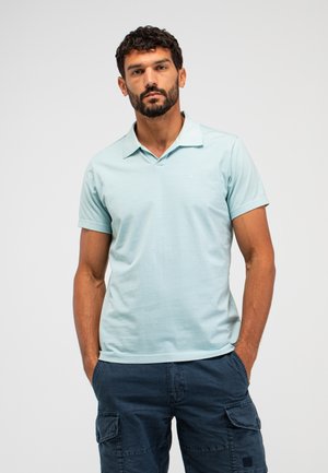 Mann mit kurzem lockigem Haar und Bart, trägt ein hellblaues Polo-Shirt mit kurzen Ärmeln und dunkelblaue Cargo-Hose, Hände in den Taschen, neutraler Hintergrund.