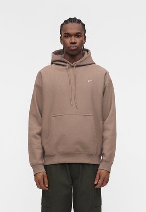 Joven con trenzas, vestido con una sudadera marrón de Nike y pantalones cargo oscuros, de pie frente a un fondo claro y liso.