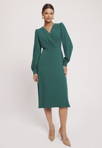 Zielona sukienka o długości midi z długimi, wyp puffowymi rękawami i dekoltem w szpic. Wykonana z gładkiego materiału, ma dopasowaną talię i zwiewny dół.