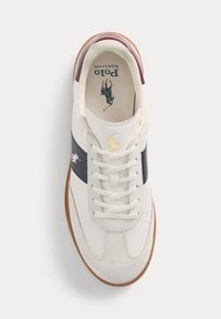 Zapatilla blanca con raya de acento en marino, suela de goma y superposiciones de ante. Presenta cordones blancos y un detalle de logo dorado en el lado.