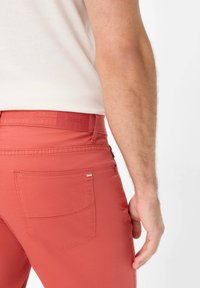 Pantaloni rossi con una texture liscia, dotati di una tasca posteriore e di una cintura con marchio. Il design presenta dettagli di cucitura minimi.