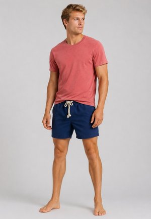 Junger Mann steht barfuß, trägt ein ausgewaschenes rotes T-Shirt und marineblaue Shorts mit weißem Kordelzug auf einem einfachen grauen Hintergrund.