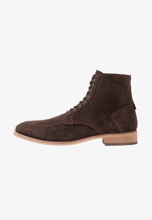 Bottine en suede marron avec un bout arrondi, semelle en cuir plate et œillets en métal pour les lacets. Elle présente des détails de couture et une languette.