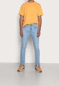 Gul bomullströja, ljusblå denimjeans och beige sneakers med bruna accenter. Modellen står på en enfärgad bakgrund.