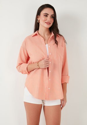Striped button-up shirt in orange and white, relaxed fit, rolled sleeves, single chest pocket, paired with white shorts. Smooth fabric texture.