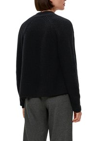 Schwarzer Strickpullover mit strukturiertem Material, Raglanärmeln und einem leicht verkürzten Rücken, kombiniert mit grauen Hosen.