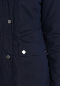 Detailní záběr na látku námořnické modré bundy s kovovými patentkami a klopovou kapsou na přední straně.
