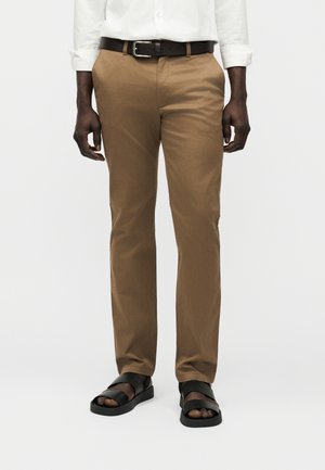 Homme portant un pantalon beige, une ceinture noire, une chemise blanche et des sandales noires à bout ouvert, debout sur un fond clair.