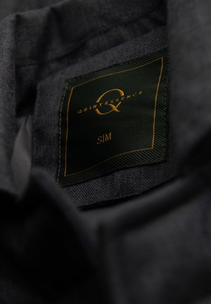 Mørkegråt stof med en sort og gylden tøjlabel, hvor der står "QUINTESSENCE Q, S/M", syet indeni.