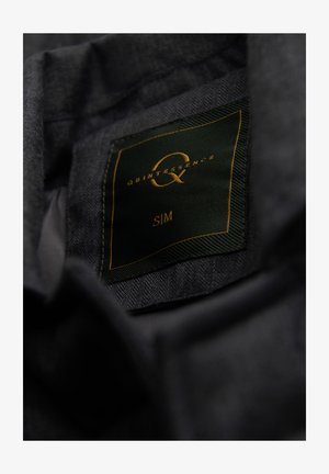 Mørkegråt stof med en sort og gylden tøjlabel, hvor der står "QUINTESSENCE Q, S/M", syet indeni.