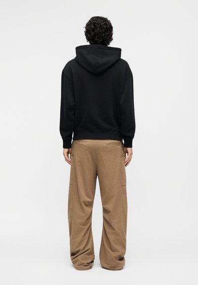 Asmuo su garbanotais plaukais, dėvintis juodą gobtuvą ir laisvo pjūvio rudos spalvos kelnes, stovintis nugara priešais paprastą foną.