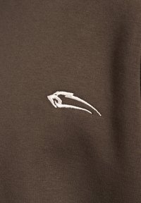 Brązowa bawełniana tkanina o teksturowanym wykończeniu, z wyraźnym białym haftowanym logo w stylizowanym kształcie po boku.