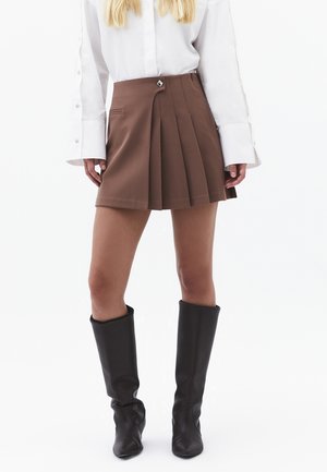Kobieta w brązowej plisowanej mini spódniczce, białej koszuli z długim rękawem i przedłużonymi mankietami oraz czarnych kozaczkach sięgających do kolan na białym tle.