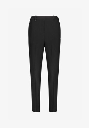 Personne marchant portant un pantalon noir slim, une chemise blanche boutonnée, un gilet sans manches en maille vert foncé et des chaussures à talons hauts noires avec des brides à chaînes.