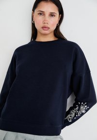 Sudadera azul marino con cuello redondo, corte holgado y intrincados bordados florales blancos en la manga izquierda. Tejido suave y texturizado.