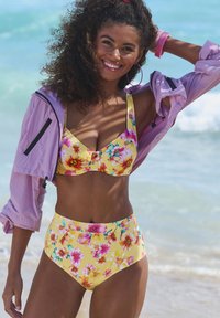Dzeltenais ziedu bikini komplekts, kas ietver bralete stila topu ar platām siksnām un augsto jostasvietu apakšdaļām, papildināts ar vieglu purpura džaku.