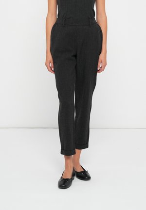 Femme portant un pantalon gris foncé taille haute, cintré, avec ourlets retroussés et des ballerines noires, debout devant un fond blanc uni.