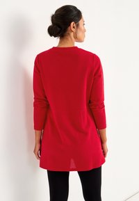 Vrouw met donker haar in een knot, gekleed in een rode langoestrik, geribbelde trui en zwarte leggings, staand tegenover een witte muur.