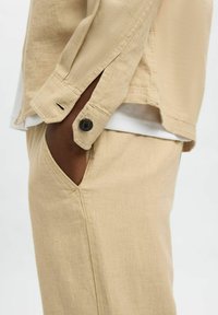 Giacca in misto cotone beige con tasca e dettagli in bottone, abbinata a pantaloni a gamba stretta coordinati, caratterizzati da una texture liscia e cuciture ordinate.