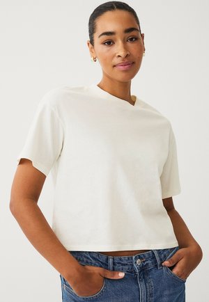 Cropped light cream T-shirt with short sleeves, round neckline, and soft texture. Worn with blue high-waisted jeans, hands in pockets.