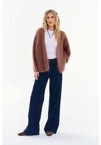 Žena stojaca, oblečená v hnedom huňatom kardigane, bielej košeli, tmavých džínsach so širokými nohavicami, topánkach s leopardím vzorom a s náhrdelníkom z korálikov na bielom pozadí.