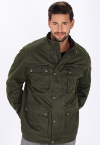 Giacca verde oliva con colletto alto, zip frontale e molteplici tasche a patta. Presenta una texture liscia e dettagli in metallo argentato.
