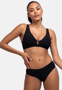 Juodas banguotas bikini komplektas su giliu iškirpimu, dvigubomis petnešomis ir atitinkančiais aukštais šortais. Lygus audinys, minimalus aksesuarų kiekis.