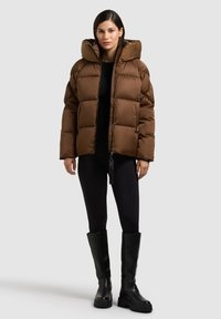 Braune Steppjacke mit Kapuze, vorne mit Reißverschluss und strukturierten Partien. Kombiniert mit schwarzen Leggings und knielangen schwarzen Stiefeln.