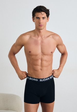 Schwarze Boxer-Shorts mit einem weißen "BJÖRN BORG"-Bund. Das Modell steht mit muskulösem Körperbau da, die Hände am Bund platziert.