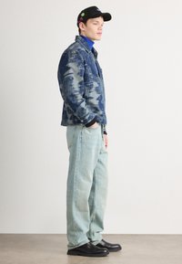 Τζάκετ από τζιν με σχέδιο σε μπλε απόχρωση, ελαφρώς ξεθωριασμένα τζιν και μαύρες ξύλινες παντόφλες. Περιλαμβάνει γιακά, τσέπες και διακοσμητική καλύπτρα.