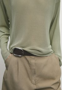 Lichtgroene, getextureerde gebreide top gecombineerd met beige plooibroek, vastgemaakt met een donkerbruine riem met een zilveren rechthoekige gesp.