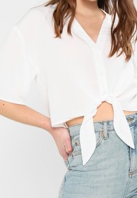 Blouse blanche courte avec un détail noué à l'avant, manches courtes, patte de boutonnage, et tissu léger, associée à un jean en denim clair taille haute.