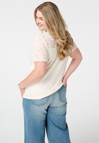 Beige top met korte kanten mouwen, geribbelde stof en een ontspannen fit. Gecombineerd met lichtblauwe wijdvallende jeans. Achteraanzicht van de outfit.