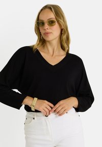 Jersey negro de cuello en V con mangas largas, combinado con pantalones blancos. La modelo también lleva gafas de sol verde claro y una pulsera dorada.