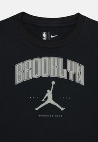 Melns kokvilnas T-krekls ar lielu pelēku tekstu "BROOKLYN", basketbolista siluetu un "EST 2012 BROOKLYN NETS" zem tā.