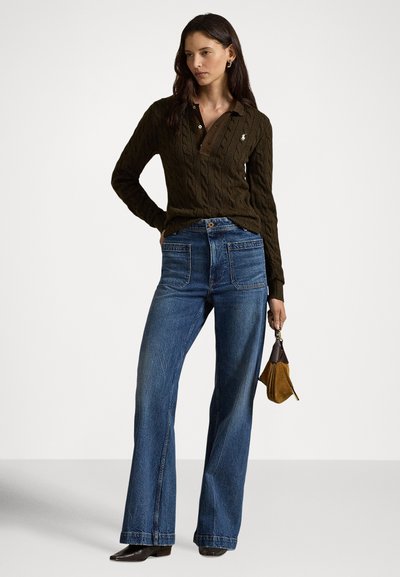 Γυναίκα που φοράει σκούρο καφέ πουλόβερ με πλέξη καναλιού και καμπάνα μπλε τζιν, κρατώντας μια μικρή καφέ τσάντα από σουέντ, μπροστά σε απλό φόντο.