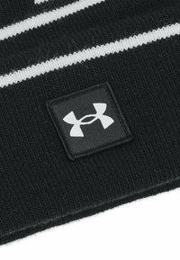 Beanie nera a maglia con strisce bianche e un'etichetta quadrata. L'etichetta presenta un logo grigio di Under Armour su uno sfondo nero. Tessuto morbido.